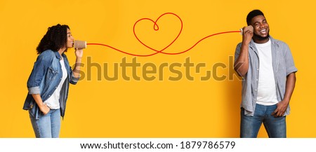 Love message. Loving african american woman telling romantic words to boyfriend through tin can phone with heart shaped string, standing together over yellow background, creative collage, panorama Royalty-Free Stock Photo #1879786579