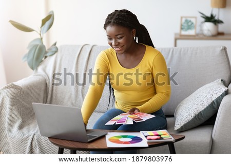 Beautiful black woman interior designer holding color swatch samples and working on laptop at home, checking palette for new design project, sitting on sofa in living room, choosing gamma for overhaul