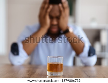Selective focus on glass of alcohol whiskey over drunk african american office manager or businessman in formal clothes holding his head. Stressed businessman, alcohol addiction concept