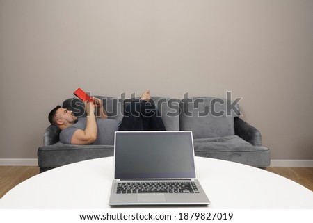 Bearded man reading red book on the couch. laptop with empty screen on white table. empty copy space for inscription. home interior.