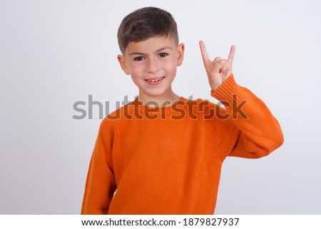 Cute Caucasian kid boy wearing knitted sweater against white wall doing a rock gesture and smiling to the camera. Ready to go to her favorite band concert.