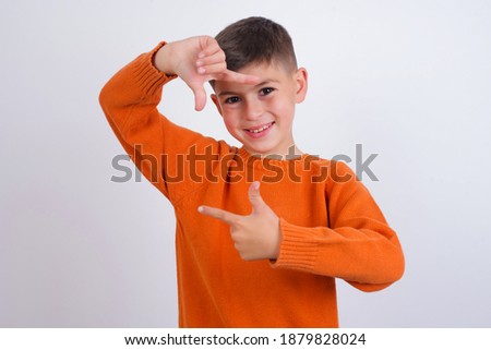 Cute Caucasian kid boy wearing knitted sweater against white wall making finger frame with hands. Creativity and photography concept.