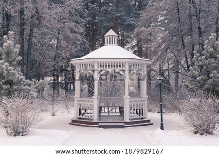 Beautiful white wooden gazebo in a winter park against the backdrop of snowy trees. Park within the city.  Royalty-Free Stock Photo #1879829167