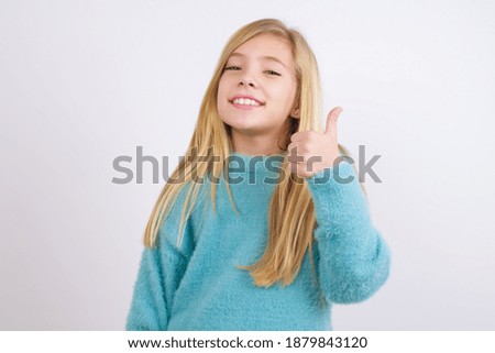Cute Caucasian kid girl wearing blue knitted sweater against white wall giving thumb up gesture, good Job! Positive human emotion facial expression body language.