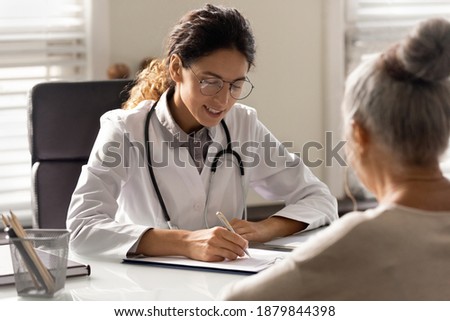 Smiling young woman GP write prescription to mature patient at consultation in hospital. Female doctor consult talk with elderly client in private clinic. Healthcare insurance, geriatrics concept. Royalty-Free Stock Photo #1879844398