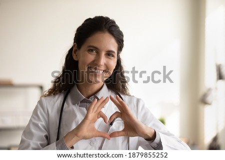 Close up portrait of smiling young Caucasian female nurse or GP in white medical uniform show heart love hand gesture. Happy woman doctor show support and care to patients or client in hospital.