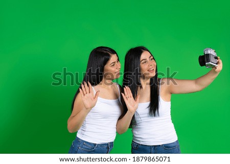 The brunette twins are smiling taking a selfie photo with a vintage camera. Green background.