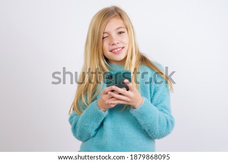 Pleased Cute Caucasian kid girl wearing blue knitted sweater against white wall using self phone and looking and winking at the camera. Flirt and coquettish concept.