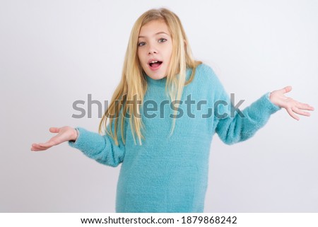 So what? Portrait of arrogant Cute Caucasian kid girl wearing blue knitted sweater against white wall shrugging hands sideways smiling gasping indifferent, telling something obvious. Royalty-Free Stock Photo #1879868242