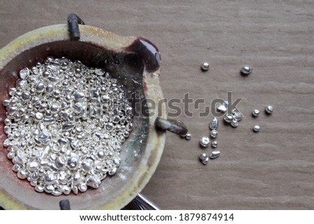 A ceramic crucible or melting pot with grains of sterling silver 925 for jewelry making after hand smelting in workshop. One of the two major precious metals on the market. Separate granules on table. Royalty-Free Stock Photo #1879874914