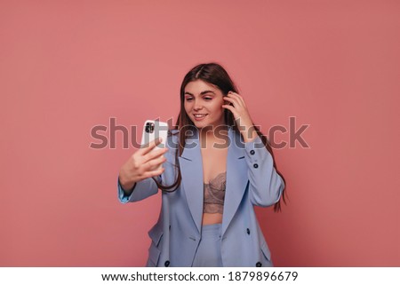 A smiling girl in a blue suit makes a selfie on a pink background. 