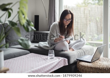 Asian woman wearing glasses writes on her notepad in front of her laptop computer while working from home sitting on a sofa in the living room of a house in Edinburgh, Scotland, United Kingdom