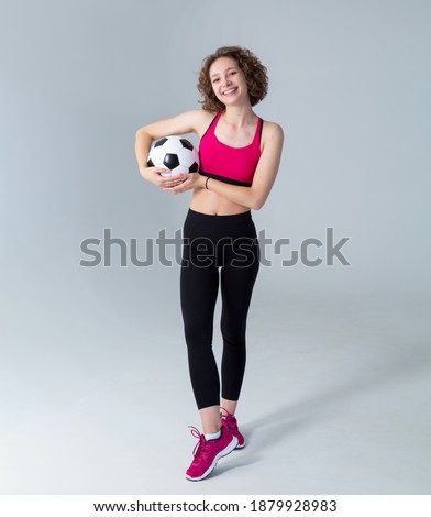 Young sportive woman with a soccer ball standing in full growth on a gray background and looking at the camera.