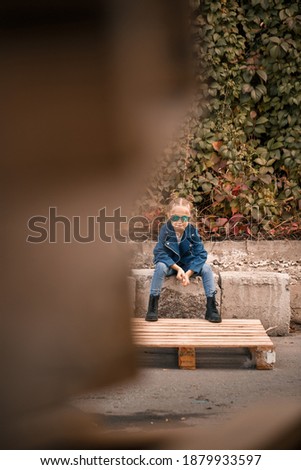 A cute little girl is sitting in sunglasses on a stone fence. Stylish child in a blue jacket. Casual style, fashion for children, fashionable suit, happy childhood
