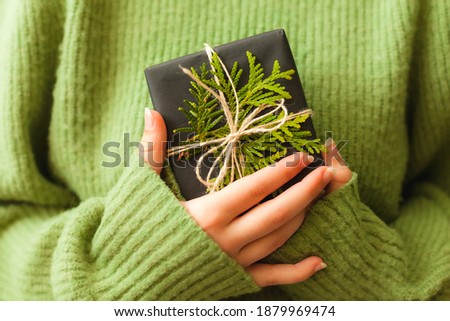 Womans hands holding nice present wrapped in black paper and decorated with green branch, close up