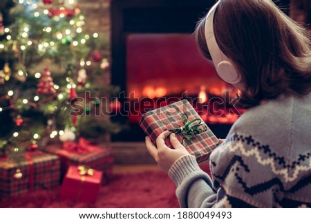 Woman in headphones with christmas gift box in hand sitting on fluffy plaid near fireplace and christmas tree. 