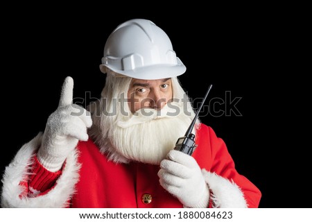 Santa Claus in construction helmet using walkie-talkie talking and pointing up on isolated black background, christmas industry concept. Royalty-Free Stock Photo #1880084623