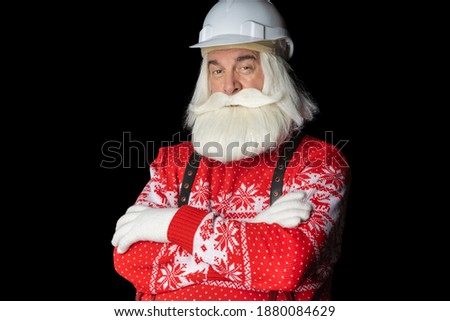 A gray-haired man in a Santa sweater with a gray beard and a helmet, his hands folded, looks slyly. Construction concept about Santa Claus Christmas Eve. Royalty-Free Stock Photo #1880084629