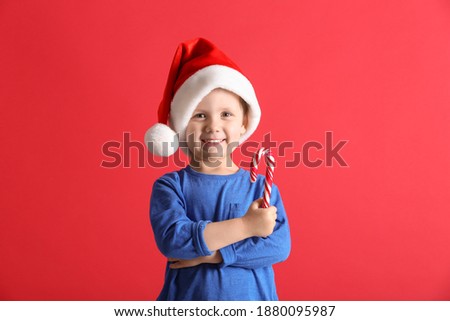 Cute little boy in Santa Claus hat holding candy cane on red background