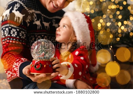 Father and daughter with snow globe near Christmas tree, focus on toy. Bokeh effect