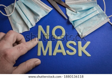 Human hand pointing the words No Mask written with plastic letters on blue paper background with a cut protective mask and scissors, concept