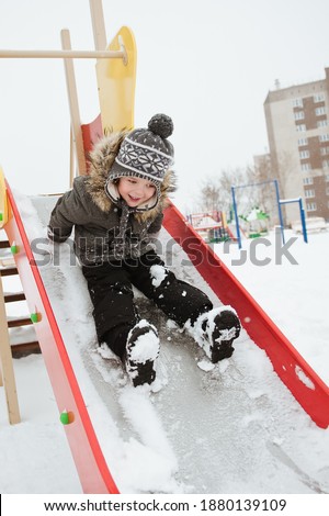 A child rides a slide on the Playground in winter. A boy in a warm coat plays in a snowy winter Park.