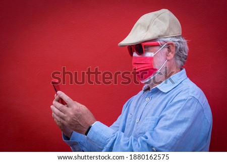 Coronavirus. Senior white haired man wearing face mask due to coronavirus looking at smart phone, standing against red background