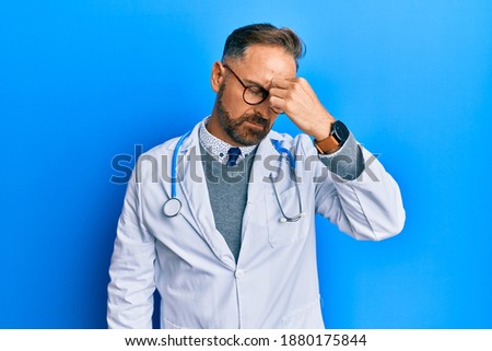 Handsome middle age man wearing doctor uniform and stethoscope tired rubbing nose and eyes feeling fatigue and headache. stress and frustration concept. 