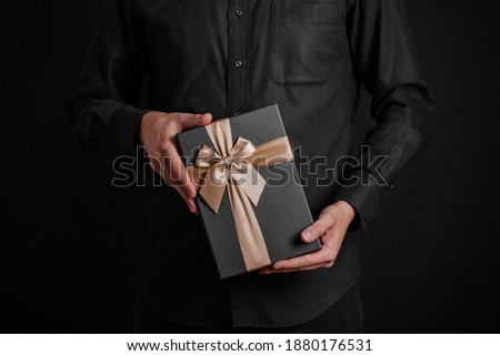 A guy in a black shirt is holding a gift with a golden ribbon. Black background. Black gift box in hands.