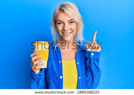 Young blonde girl holding take away coffee smiling with an idea or question pointing finger with happy face, number one 