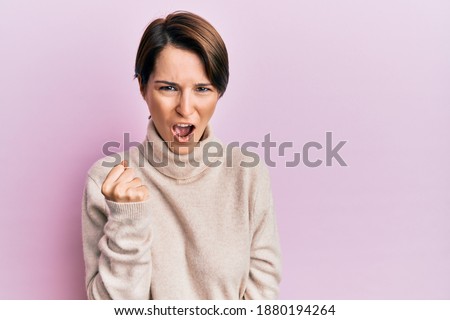 Young brunette woman with short hair wearing casual winter sweater angry and mad raising fist frustrated and furious while shouting with anger. rage and aggressive concept. 