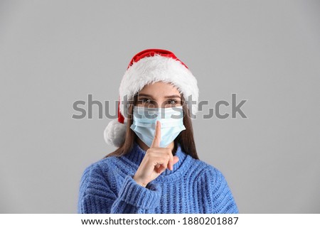 Pretty woman in Santa hat and medical mask showing silence gesture on grey background