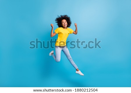 Full size photo of afro american trendy brunette little girl jump win wear yellow t-shirt jeans isolated on blue color background