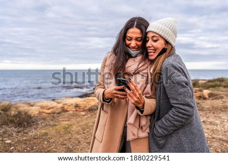 Young mixed race female LGBTQ couple having fun making faces with tongue out laughing watching photos on smartphone - Two beautiful lesbian women joking with internet outdoor in sea winter vacation