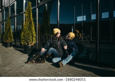 Portrait of a beautiful young happy couple man and woman in yellow hats and warm jackets sitting, hugging near modern glass architecture. Girlfriend, style, boyfriend, office house.