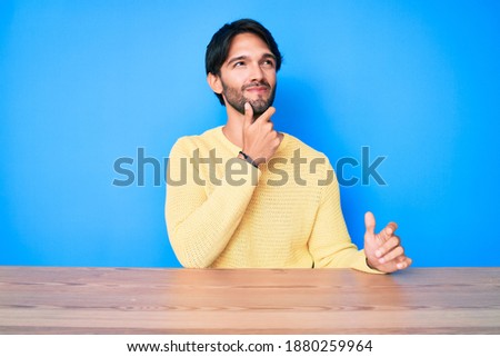 Handsome hispanic man wearing casual sweater sitting on the table serious face thinking about question with hand on chin, thoughtful about confusing idea 