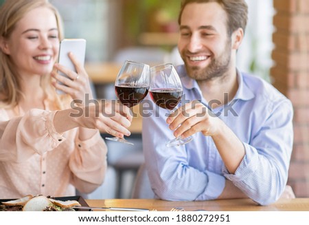 Young woman making photo of romantic toast with clinking wine glasses during festive dinner at cafe, selective focus. Affectionate couple celebrating Valentine's Day or anniversary
