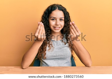 Teenager hispanic girl wearing casual clothes sitting on the table doing money gesture with hands, asking for salary payment, millionaire business 