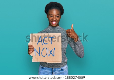 Young african american girl holding act now banner smiling happy and positive, thumb up doing excellent and approval sign 