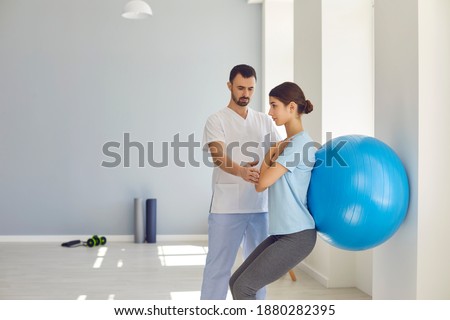 Physiotherapist helping young woman do wall squats with fit ball to get rid of backache and regain spinal health. Young female patient doing back exercise using fitball in physio room of modern clinic Royalty-Free Stock Photo #1880282395