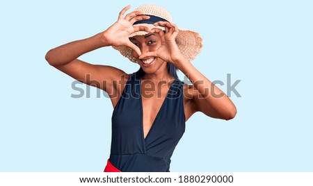 Young african american woman wearing swimsuit and summer hat doing heart shape with hand and fingers smiling looking through sign 