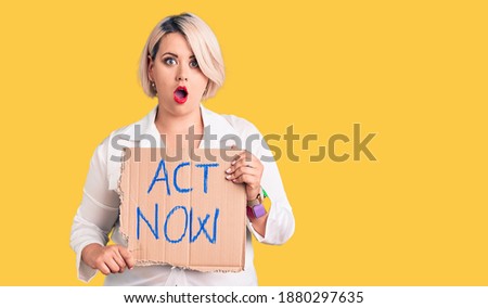 Young blonde plus size woman holding act now cardboard banner scared and amazed with open mouth for surprise, disbelief face 