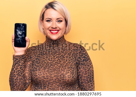 Young beautiful blonde plus size woman holding broken smartphone showing cracked screen looking positive and happy standing and smiling with a confident smile showing teeth