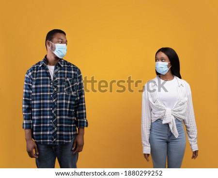 Relationships and social distancing during covid-19 lockdown. Serious young african american students guy and lady in protective masks looking at each other, isolated on yellow background, empty space