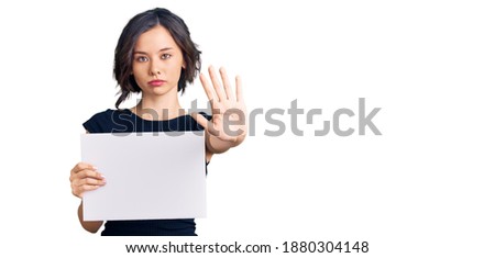 Young beautiful girl holding blank empty banner with open hand doing stop sign with serious and confident expression, defense gesture 