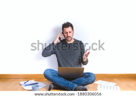 attractive man on his 40s sitting on the floor of an apartment with a laptop and discussing by phone doings gestures with hand