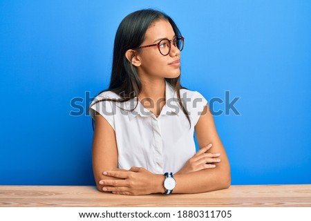 Beautiful hispanic woman wearing casual clothes sitting on the table looking to the side with arms crossed convinced and confident 