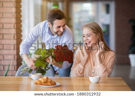 Affectionate millennial man surprising his girlfriend, giving her bouquet of flowers in cafe. Loving young couple celebrating anniversary, birthday or Valentine's Day together at canteen