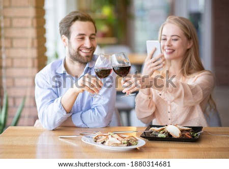 Portrait of lovely lady taking picture of toast with clinking wine glasses during holiday dinner at restaurant. Romantic millennial couple celebrating anniversary or Valentine's Day, saying cheers