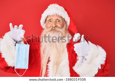 Old senior man wearing santa claus costume holding safety mask smiling happy and positive, thumb up doing excellent and approval sign 